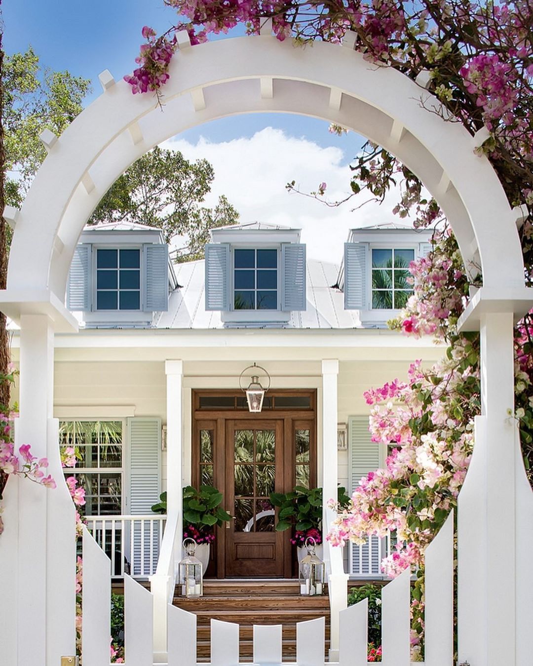 Breathtaking lovely curb appeal with amaazing floral covered arbor and blue shutters - Pineapples Design Group. Charming inspiration if you love white painted house exteriors! #whitehouses #housedesign #exteriors #curbappeal #shutters #arbor