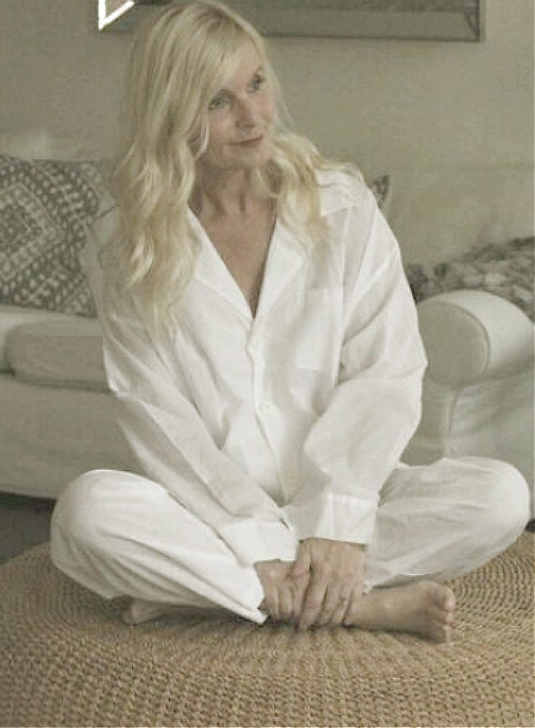 Michele of Hello Lovely in white men's style pajamas sitting on a rattan ottoman.